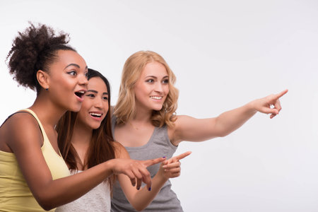 Half-length portrait of three young lovely girls wearing colorful T-shirt looking and pointing at someone with surprise. Isolated on white backgroundの写真素材