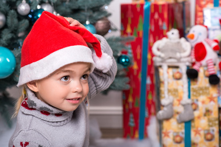 Selective focus on the little cute fair-haired smiling girl sitting on the floor near Christmas presents wearing warm sweater and red cap of Santa Claus thinking about the chocolateの写真素材