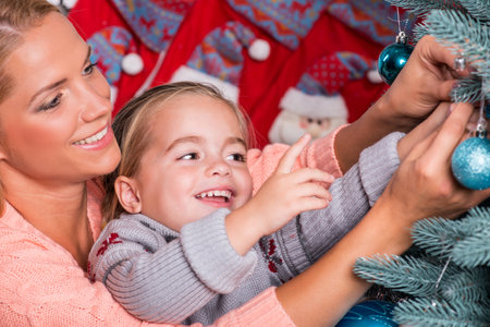 Half-length portrait of the little cute fair-haired smiling girl with her lovely mom wearing warm sweaters and jeans sitting aside near the Christmas tree decorating itの写真素材