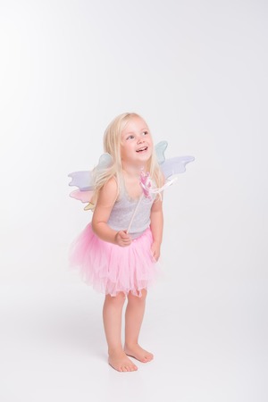 Full-length portrait of little fair-haired lovely girl wearing pretty grey vest pink skirt and white wings standing with her magic wand and laughing. Isolated on white backgroundの写真素材