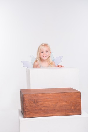 Half-length portrait of little fair-haired lovely smiling girl wearing pretty grey vest pink skirt and white wings looking out of the wooden chair. Isolated on white backgroundの写真素材