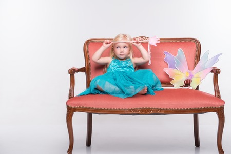 Full-length portrait of little fair-haired lovely girl wearing pretty blue dress sitting on the wonderful sofa near the white wings thinking about something. Isolated on white backgroundの写真素材