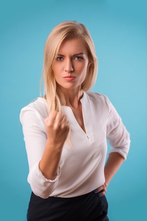 Half-length portrait of beautiful angry blonde wearing white classic blouse and black skirt standing threatening us. Isolated on blue backgroundの写真素材