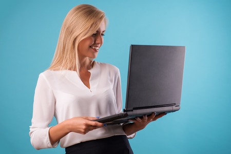 Half-length portrait of beautiful smiling business lady wearing white classic blouse and black skirt standing aside looking for something in her laptop. Isolated on blue backgroundの写真素材