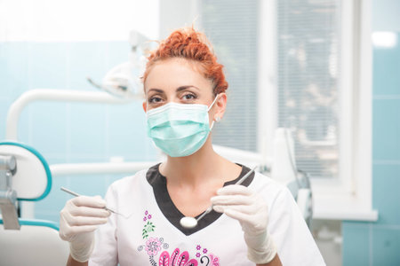 Half-length portrait of young dentist wearing medical mask and gloves standing holding tweezers and little mirror wanted to examine her patientの写真素材