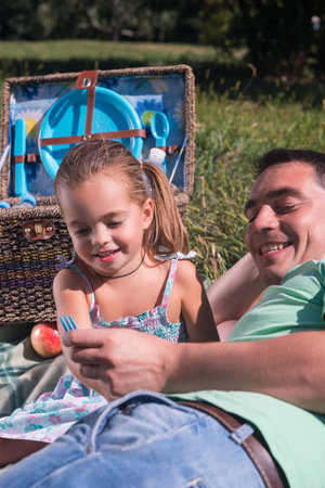 Selective focus on happy father lying on the plaid with his lovely smiling daughter talking about something near the wicker basket standing on backgroundの写真素材