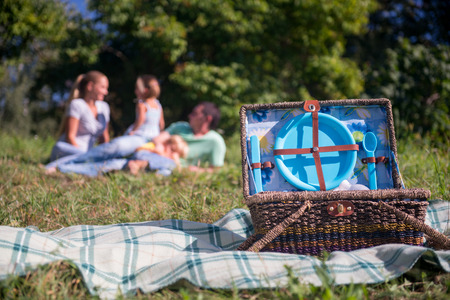 Selective focus on the great wicker basket for picnic standing on the plaid on grass with blue crockery in it. Happy family lying on backgroundの写真素材