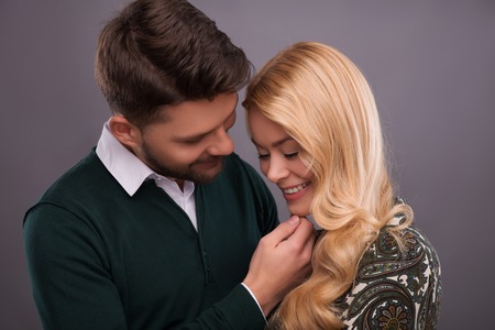 Half-length portrait of handsome young man wearing white shirt and dark-green cardigan standing hugging his beautiful fair-haired girlfriend wanted to kiss herの写真素材