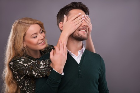 Half-length portrait of beautiful smiling blonde standing behind her handsome boyfriend closing his...