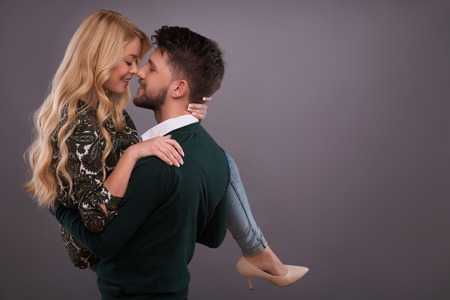 Half-length portrait of handsome smiling man wearing white shirt and green jumper standing aside raising up her beautiful happy girlfriend looking at her. Isolated on dark backgroundの写真素材