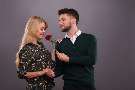 Half-length portrait of young handsome man wearing green sweater and white shirt hugging and presenting his lovely fair-haired smiling girlfriend wonderful red rose. Isolated on grey backgroundの写真素材