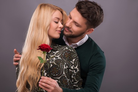 Half-length portrait of young handsome man wearing green sweater and white shirt hugging and presenting his lovely fair-haired smiling girlfriend wonderful red rose. Isolated on grey backgroundの写真素材