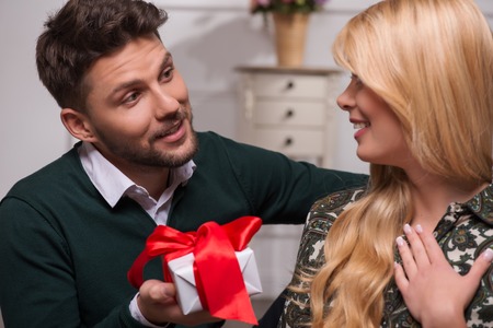 Selective focus on the beautiful surprised fair-haired young woman wearing blouse and jeans looking happy at her handsome boyfriend giving her wonderful presentの写真素材