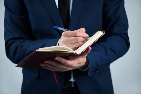 Selective focus on the dairy in the hands of busy man wearing white shirt tie and blue jacket writing something in itの写真素材