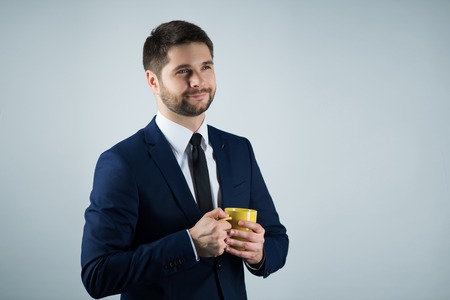 Half-length portrait of handsome young smiling man wearing white shirt tie and blue suit holding a yellow cup of coffee exulting in the morning. Isolated on white backgroundの写真素材