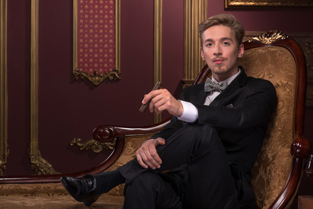 Half-length portrait of handsome young fair-haired man wearing classic costume sitting relaxing on the comfortable chair with his favorite cigar satisfied with his lifeの写真素材