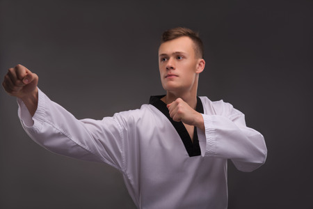 Half-length portrait of young handsome fair-haired karate enthusiast wearing white kimono coaching himself before the important fight. Isolated on the dark backgroundの写真素材