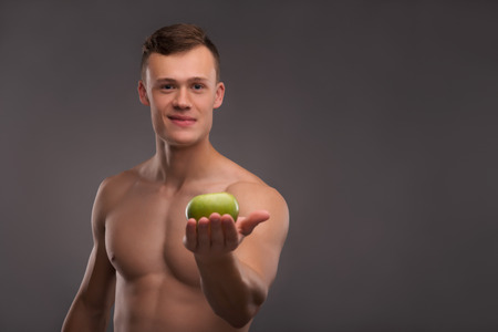 Selective focus on the young handsome fair-haired bare-chested karate enthusiast holding on his palm delicious green apple on backgroundの写真素材