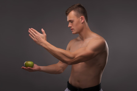 Half-length portrait of young handsome fair-haired bare-chested karate enthusiast standing aside training his attention focusing on the apple. Isolated on the dark backgroundの写真素材