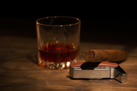 Selective focus on the Cuban cigar lying on the lighter near the glass of whiskey standing on the wooden table on backgroundの写真素材