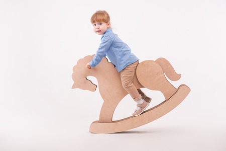 Full-length portrait of little lovely smiling girl wearing blue shirt and brown pants swaying on the wooden toy horse. Isolated on the white backgroundの写真素材