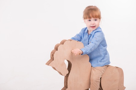 Full-length portrait of little lovely smiling girl wearing blue shirt and brown pants swaying on the wooden toy horse. Isolated on the white backgroundの写真素材