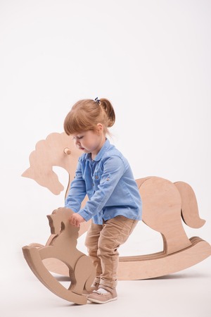 Full-length portrait of little lovely girl wearing blue shirt and brown pants standing near the wooden toy horse playing with the little one. Isolated on the white backgroundの写真素材