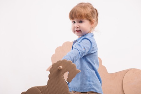 Selective focus on the little lovely girl wearing blue shirt and brown pants playing with the little wooden toy horse. Bigger one standing on backgroundの写真素材