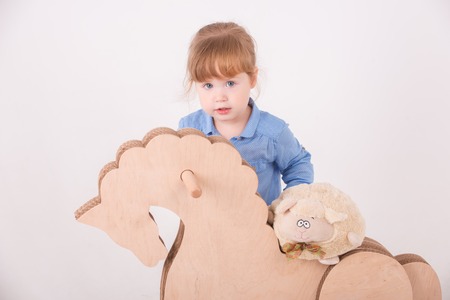 Half-length portrait of little lovely girl wearing blue shirt and brown pants leaning on the wooden horse holding a soft toy. Isolated on the white backgroundの写真素材