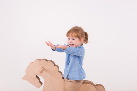Half-length portrait of little lovely smiling girl wearing blue shirt and brown pants riding on the wooden horse. Isolated on the white backgroundの写真素材