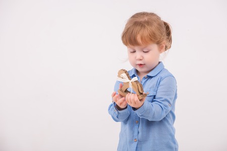 Half-length portrait of little lovely smiling girl wearing blue shirt and brown pants holding in her hands little wooden horse. Isolated on the white backgroundの写真素材