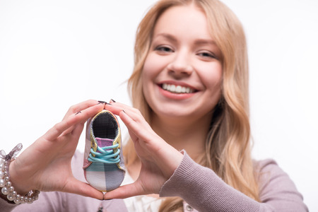 Portrait of young attractive happy Caucasian woman with long fair hair smiling and holding baby shoes, selective focus isolated on white background, baby expectation conceptの写真素材
