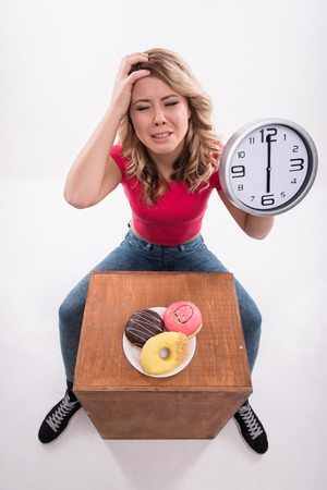 Time for diet slimming. Young beautiful woman with clock keeps from eating doughnuts after 6 pm - weight loss concept, top view isolated on a white backgroundの写真素材