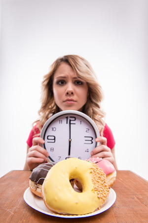 Time for diet slimming. Young beautiful woman with clock keeps from eating doughnuts after 6 pm - weight loss concept, close-up portrait selected focus isolated on a white backgroundの写真素材