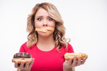 Half-length portrait of beautiful young scared blonde wearing pink T-shirt standing with the tied tape measure on her mouth cannot taste tasty doughnuts in her hands. Isolated on white backgroundの写真素材