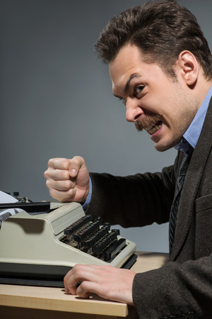 Lost inspiration. Side view of young nerd author striking his typewriter while sitting at his working place against grey backgroundの写真素材
