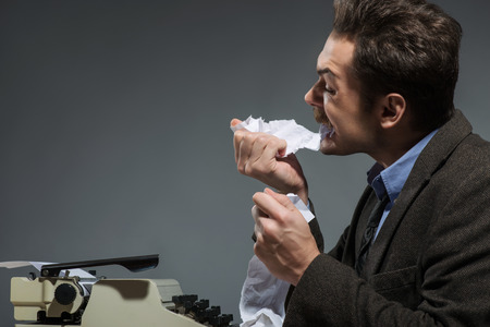 Waiting for idea. Side view of excited young author working at the typewriter and tearing paper while sitting at his working place against grey backgroundの写真素材