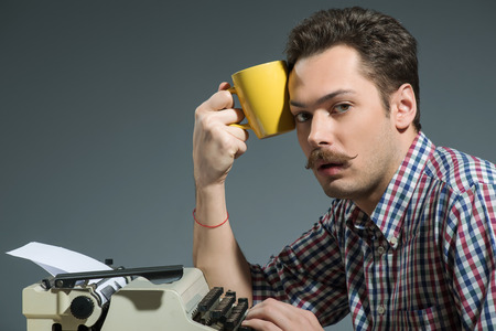 Sleepless night. Creative young author working at the typewriter with cup of coffee while sitting at his working place against grey backgroundの写真素材