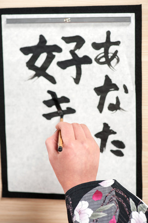 Traditional calligraphy writing. Top view image of woman hand in kimono writing Japanese or Chinese hieroglyphs on paper arranged on wooden tableの写真素材