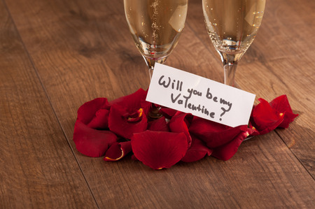 Let us celebrate. Closeup image of champagne glasses decorated with rose petals and greeting card devoted to Saint Valentines Day celebration arranged on wooden backgroundの写真素材