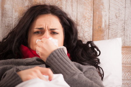 Sick woman caught a cold. Closeup image of frustrated young woman with red nose lying in bed with thick scarf and sneezing into tissueの写真素材