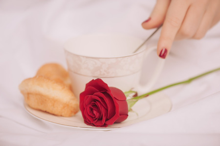 Morning coffee. Closeup image of red rose in selective focus and a cup of coffee with croissantの写真素材