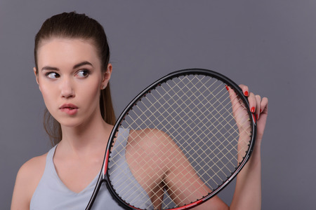 Not sure of victory. Closeup portrait of beautiful young woman in sports clothes holding tennis racket and biting lips while standing against white background with copy spaceの写真素材
