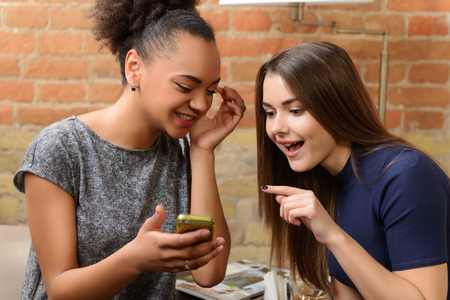Happy friends in restaurant. Two young women having fun and watching photos on cell phone while drinking coffee at the restaurantの写真素材