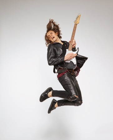For those who know how to rock. Full-length image of handsome young guitar player in leather suit jumping with the guitar and singing isolated on grey backgroundの写真素材