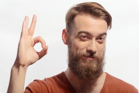 Everything is OK. Portrait of handsome tattooed bearded man wearing orange tshirt and smiling away showing OK sign while standing against white backgroundの写真素材