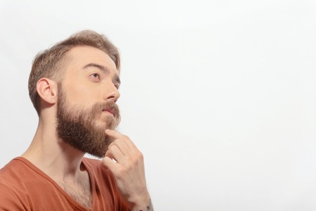 Deep in thoughts. Young bearded man touching his chin with a finger looking at copy space while...