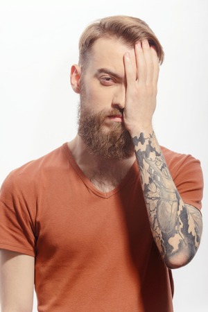 Morning after the party.  Young bearded man covering his face with one palm looking frustrated while standing against white backgroundの写真素材