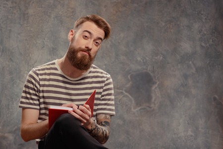 Young and creatively independent.  Portrait of young bearded tattooed man in a striped longsleeve looking at camera and keeping his notepad and pencil while sitting against dark backgroundの写真素材