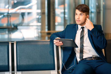 Always on the go. Portrait of handsome smiling businessman in formalwear holding a digital tablet with earphones while sitting in the airport in business loungeの写真素材